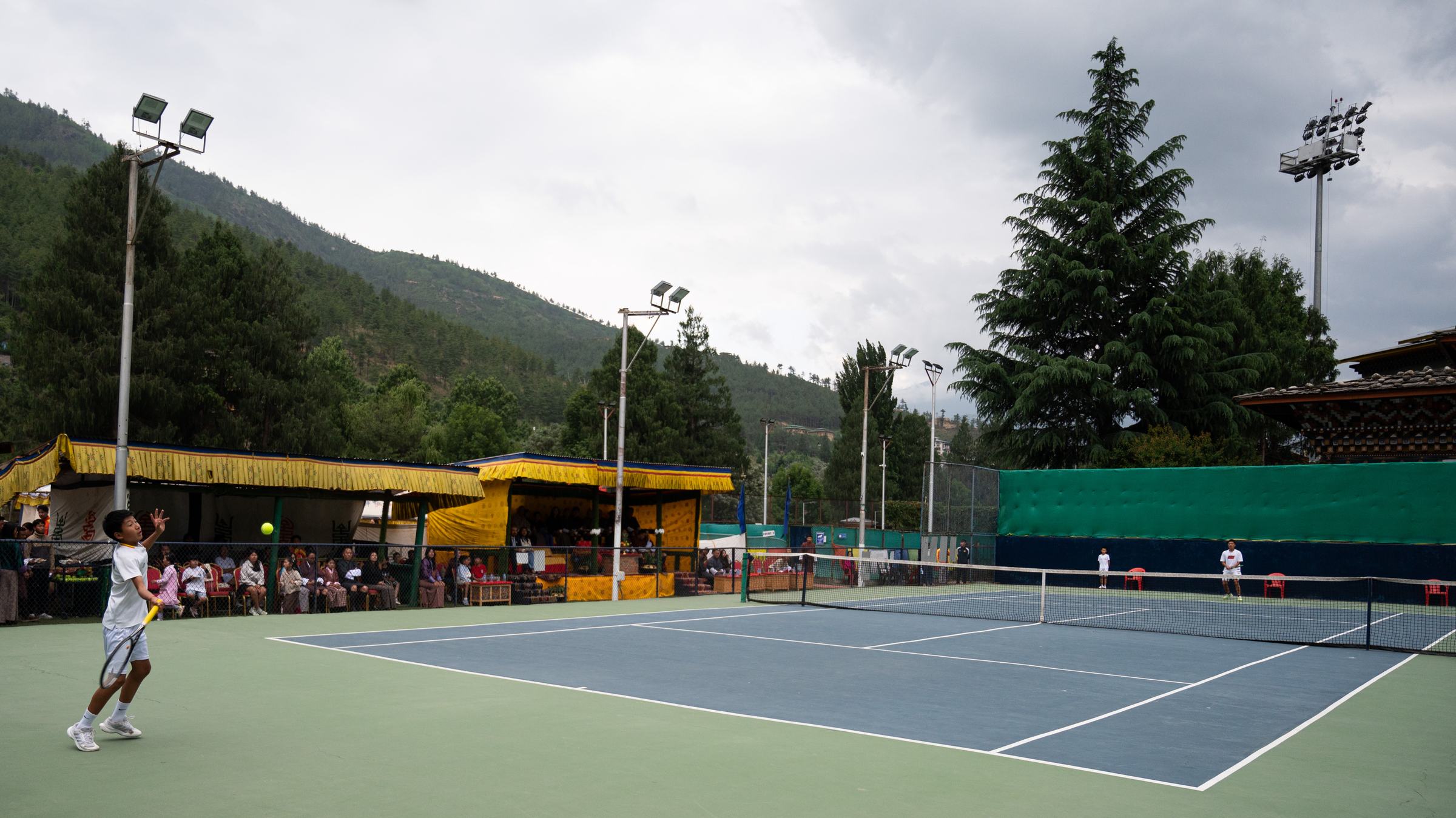 A junior player serving during the U16 final at Changlimithang, spectators under yellow canopies and forested Bhutanese hills behind